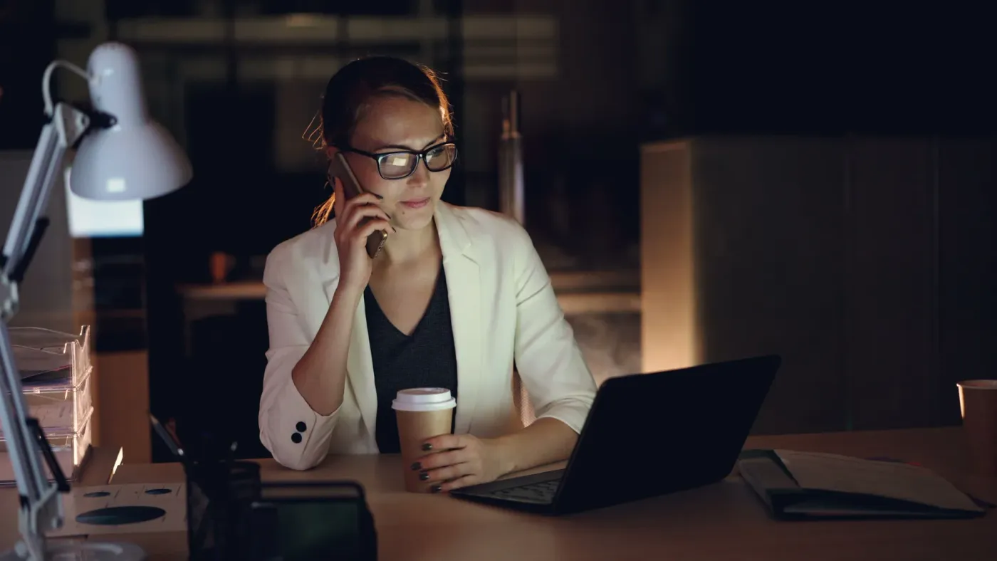 A professional on the phone at her desk at night, reviewing an application on her laptop